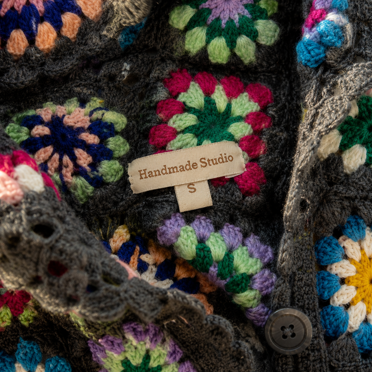 Close-up of a handmade crocheted cardigan featuring multicolor granny square motifs on a dark charcoal gray base. A small fabric tag reading 'Handmade Studio' and 'S' is visible, along with a dark gray button.