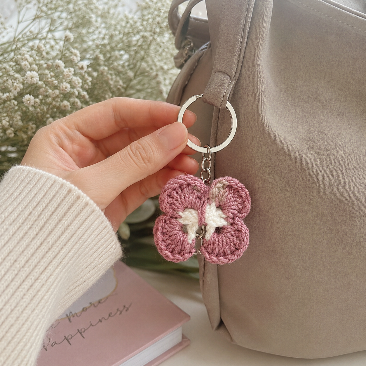 A hand holds a pink crocheted flower keychain with a silver metal ring, positioned next to a taupe-colored handbag with a book in the background.